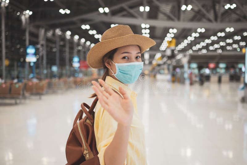 Asian woman backpacker wearing protective face mask stand and raise hand to say goodbye in an international airport