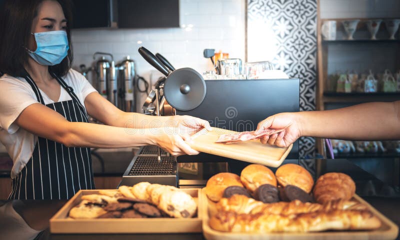 Asian woman, Coffee shop business owner wearing a surgical mask, accept cash payments from customers, avoiding contact By placing money on a wooden tray, to coffee shop and preventing the spread of virus concept