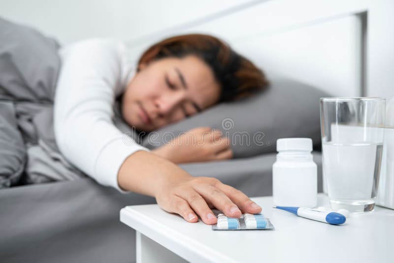 Asian woman feeling sick and using hands to touching pills on the table while lying in blanket on the bed