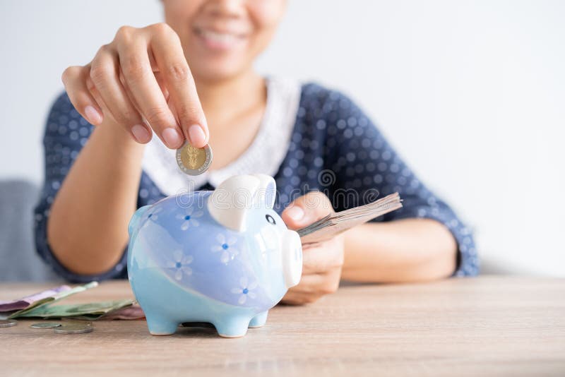 Asian woman hand is putting a coin in a piggy bank and banknote on the table at home, Investment business and retirement. finance. And saving money for the stock photos Asian woman hand is putting a coin in a piggy bank and banknote on the table at home, Investment business and retirement. finance. And saving money for the stock photos