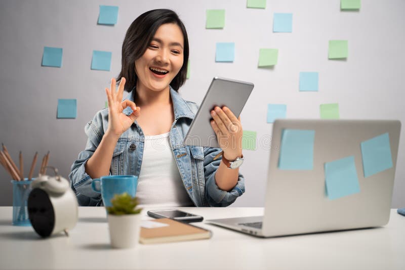 Asian woman happy looking at tablet reading news showing OK sign at home office. WFH. Work from home. Prevention Coronavirus COVID. 19 concept royalty free stock images