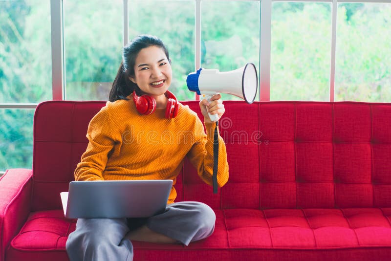 Asian woman holding megaphone with headphone and computer,sitting on red sofa in the living room of  house,working at home,with