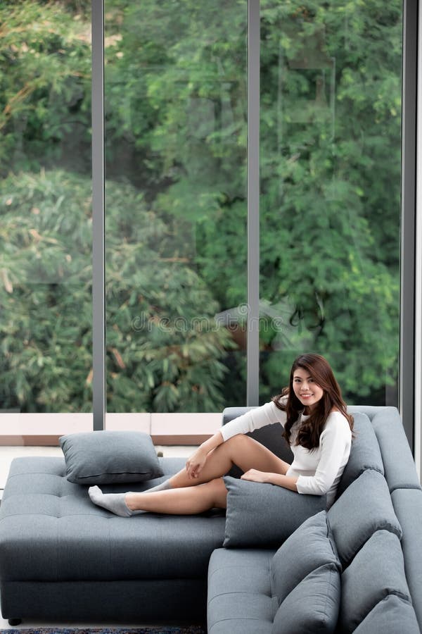 Asian woman sitting on sofa near big glass windows, relaxing alone in house with green forest in background