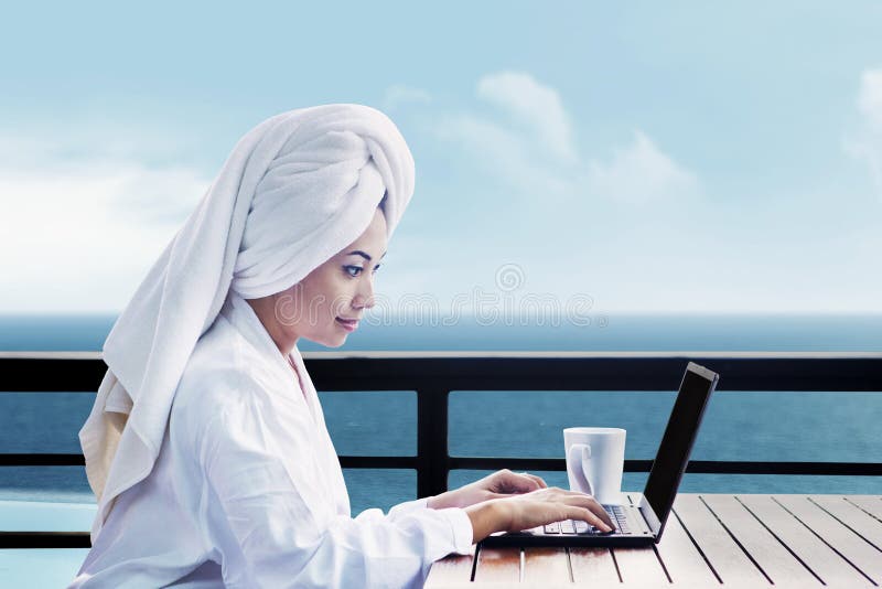 Asian woman in bathrobe with mug of coffee and typing on laptop computer. shot on ocean view. Asian woman in bathrobe with mug of coffee and typing on laptop computer. shot on ocean view