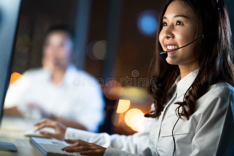 Asian woman work as customer support service or call center phone operator, using desktop computer and microphone headset, late night shift. Overtime office life, telemarketing or sales job concept. Asian woman work as customer support service or call center phone operator, using desktop computer and microphone headset, late night shift. Overtime office life, telemarketing or sales job concept