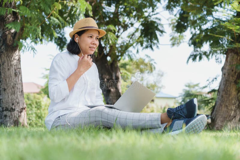 Asian women wear hat are using laptop with wired headphones while sitting on grass green In the public park stock photography Asian women wear hat are using laptop with wired headphones while sitting on grass green In the public park stock photography
