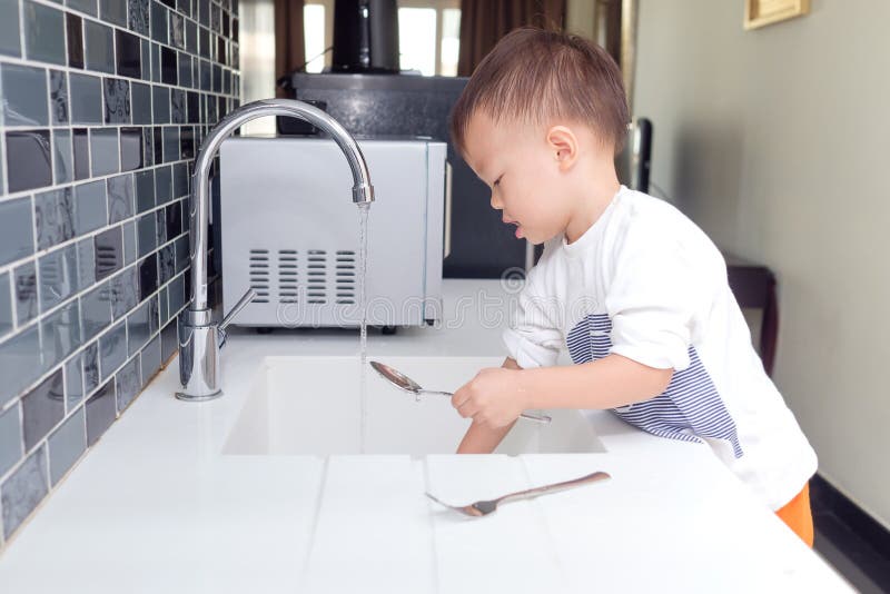 Asian 2 year old toddler boy child standing & having fun doing the dishes, concentrate on washing dishes in kitchen at home