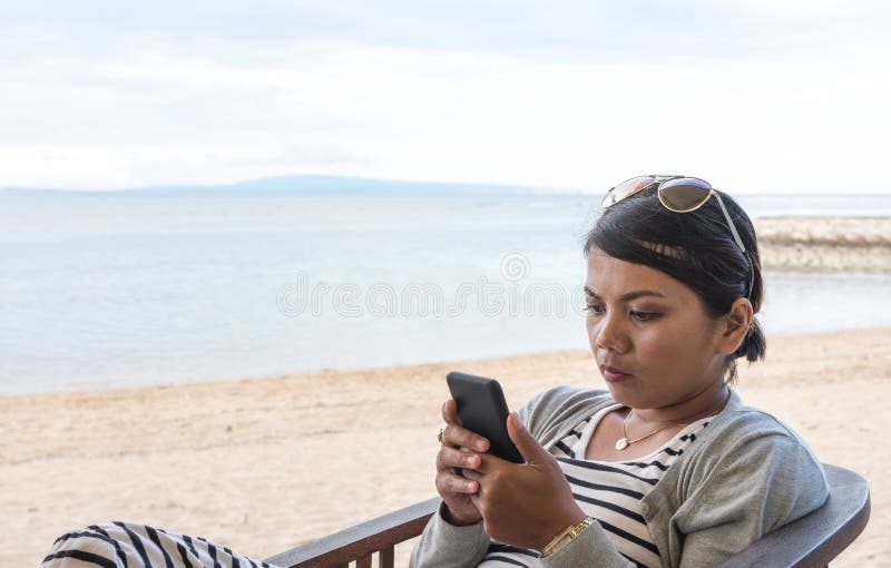 Asiatische Frau, die am Strand sitzt und ihr Handy beim Lesen anschaut lizenzfreies stockfoto Asiatische Frau, die am Strand sitzt und ihr Handy beim Lesen anschaut lizenzfreies stockfoto
