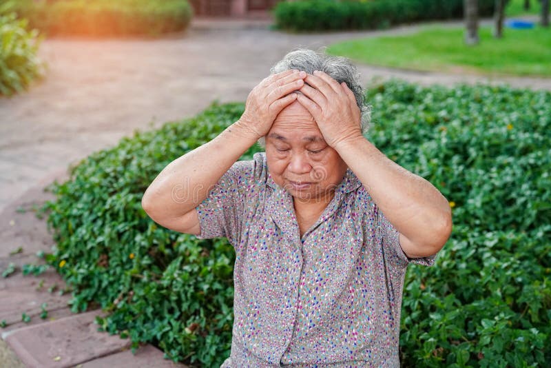 Asiatische geduldige Kopfschmerzen älterer oder älterer Frau alter Dame beim Sitzen auf Bett in Pflegekrankenstation
