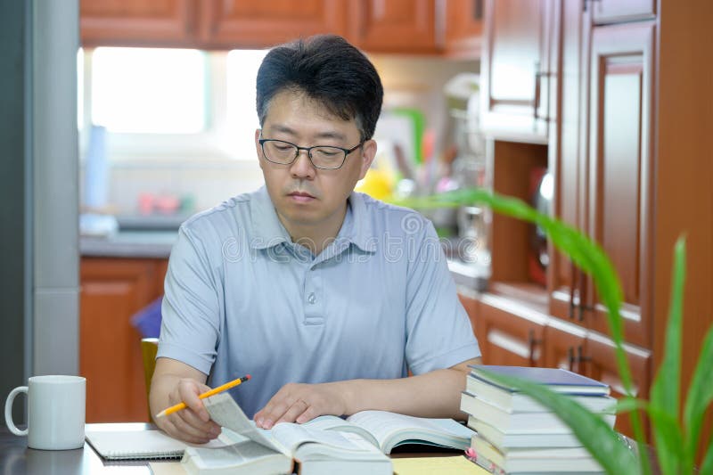 Asiatischer Mann im mittleren Alter, der zu Hause am Schreibtisch liest ein Buch und ein Studium sitzt.