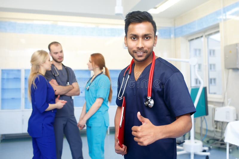 Asiatischer männlicher Doktor vor dem Team, Kamera oben betrachtend, Showdaumen, mit Ärzteteam im Hintergrund Gemischtrassiges Te stockfoto Asiatischer männlicher Doktor vor dem Team, Kamera oben betrachtend, Showdaumen, mit Ärzteteam im Hintergrund Gemischtrassiges Te stockfoto