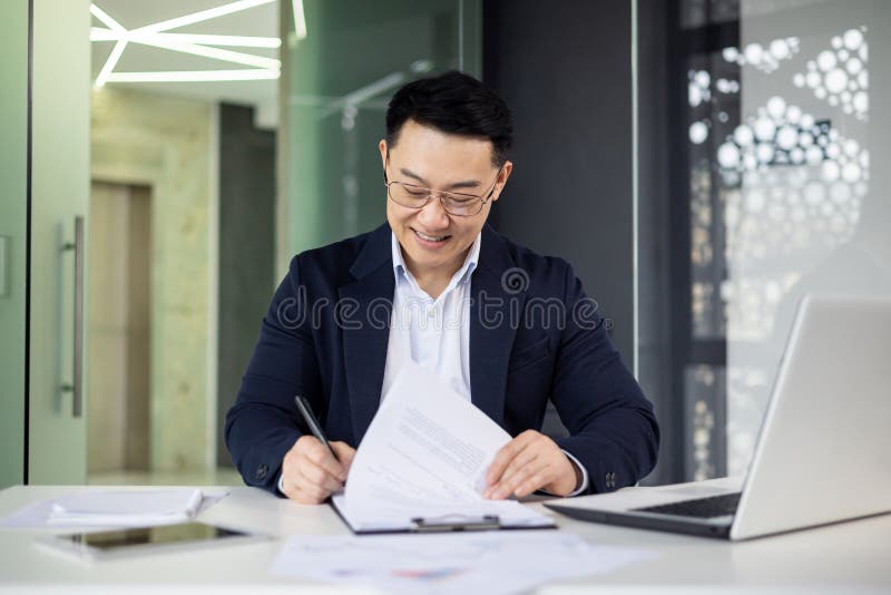 Smiling Asian businessman in a formal suit reviews documents while working on a laptop in a contemporary office setting with natural lighting. Smiling Asian businessman in a formal suit reviews documents while working on a laptop in a contemporary office setting with natural lighting.