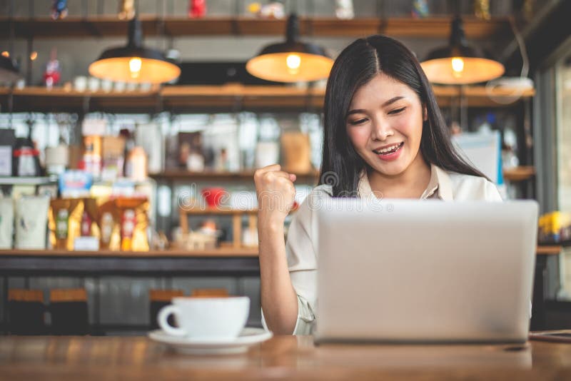 Asian female freelancer doing happy gesture by raising hand when using laptop in cafe. Business and success concept. Coffee shop and outdoors theme. Cafeteria background