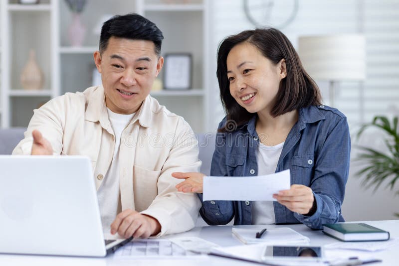 Asian couple engaging in household financial planning with a laptop in living room, discussing and reviewing their finances, with papers ,image captures happiness, teamwork, and digital convenience. Asian couple engaging in household financial planning with a laptop in living room, discussing and reviewing their finances, with papers ,image captures happiness, teamwork, and digital convenience.