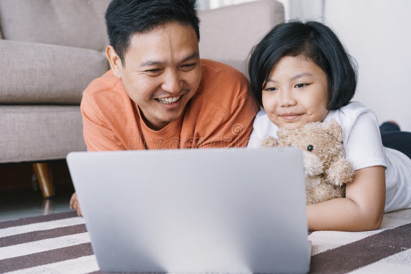 Asiens far och dotter använder en bärbar dator medan de ljuger på mattan i vardagsrummet hemma Den söta lilla flickan kramar dock royaltyfri fotografi