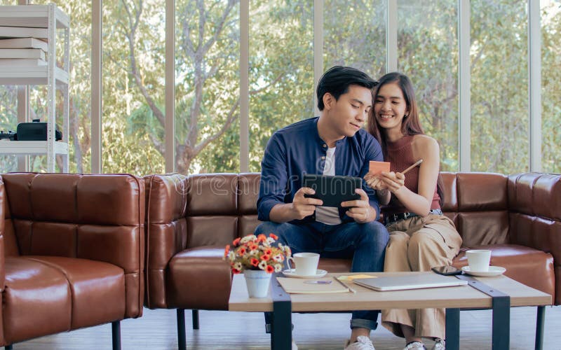 Asiática adorable dulce feliz pareja sentada en el sofá de cuero marrón en la sala de estar en casa mientras escribe el mensaje y imagen de archivo libre de regalías