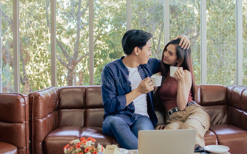 Asiática feliz pareja sentado en el sofá en la sala de estar en casa mientras bebe sosteniendo taza de café sonriendo abrazando y imagen de archivo libre de regalías