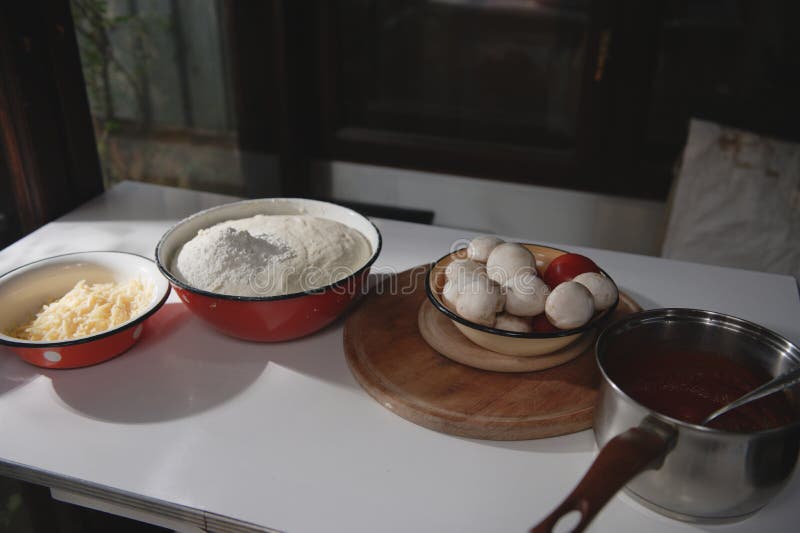 Assortment of cooking ingredients for preparing homemade pizza : rising yeast dough, grated cheese, fresh champignons and tomatoes in enamel bowls on the table in rustic kitchen. Food still life. Assortment of cooking ingredients for preparing homemade pizza : rising yeast dough, grated cheese, fresh champignons and tomatoes in enamel bowls on the table in rustic kitchen. Food still life