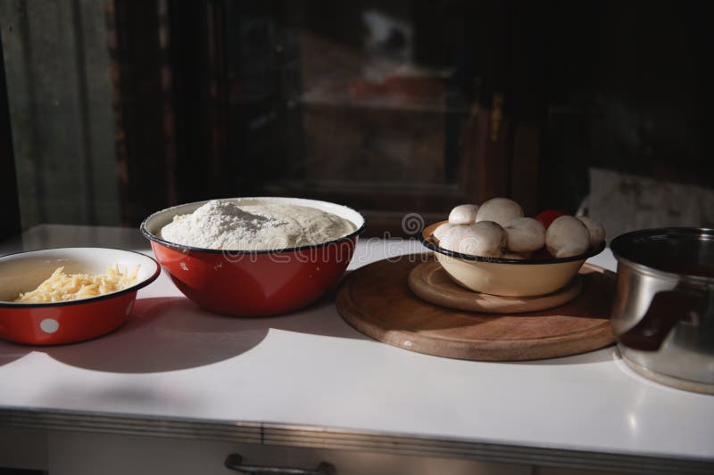 Assortment of baking ingredients for preparing homemade pizza : rising yeast dough, grated cheese, fresh champignons and tomatoes in enamel bowls on the table in rustic kitchen. Food still life. Assortment of baking ingredients for preparing homemade pizza : rising yeast dough, grated cheese, fresh champignons and tomatoes in enamel bowls on the table in rustic kitchen. Food still life