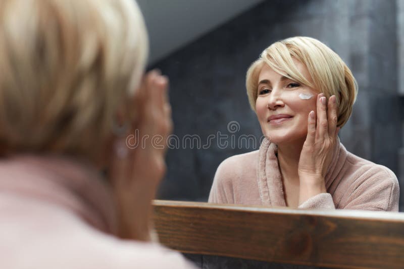 Astuzia. una donna matura usa una crema cosmetica sulla pelle di fronte al riflesso dello specchio.