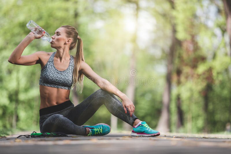 Athletic lady is sitting on road during break in work out. She is drinking water from bottle and quenching thirst. Female is having jumping rope on ground because she is using it for cardio training. Athletic lady is sitting on road during break in work out. She is drinking water from bottle and quenching thirst. Female is having jumping rope on ground because she is using it for cardio training