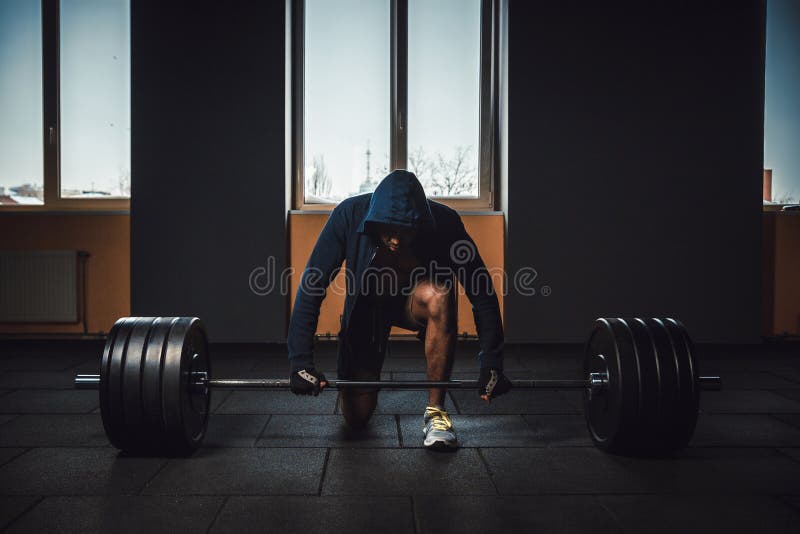 Athletic man in jacket with a hood waiting and preparing before lifting heavy barbell. fitness, sport, training, gym and lifestyle