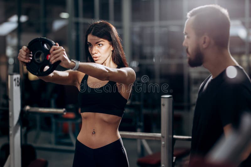 Athletisches Mädchen, das Übung für die Muskeln der Arme mit Gewicht in der Turnhalle unter Aufsicht eines Trainers tut lizenzfreie stockfotos