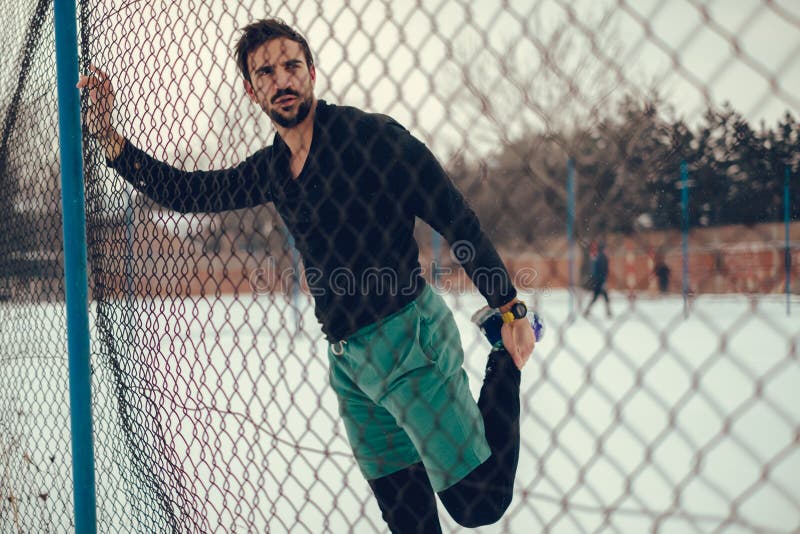 Male athlete stretching quadriceps on the fence on a snowy day. Male athlete stretching quadriceps on the fence on a snowy day