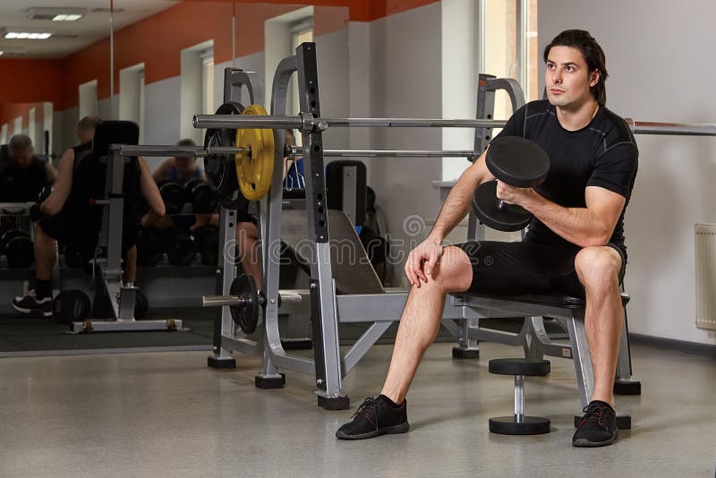 Atleta construido muscular joven que se resuelve en un gimnasio, sentándose en una máquina del levantamiento de pesas y las pesas imagen de archivo libre de regalías Atleta construido muscular joven que se resuelve en un gimnasio, sentándose en una máquina del levantamiento de pesas y las pesas imagen de archivo libre de regalías
