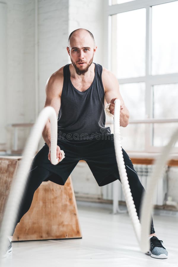 Atleta del hombre del ajuste que usa las cuerdas de la batalla para ejercitar en el pasillo de deporte ligero foto de archivo libre de regalías Atleta del hombre del ajuste que usa las cuerdas de la batalla para ejercitar en el pasillo de deporte ligero foto de archivo libre de regalías