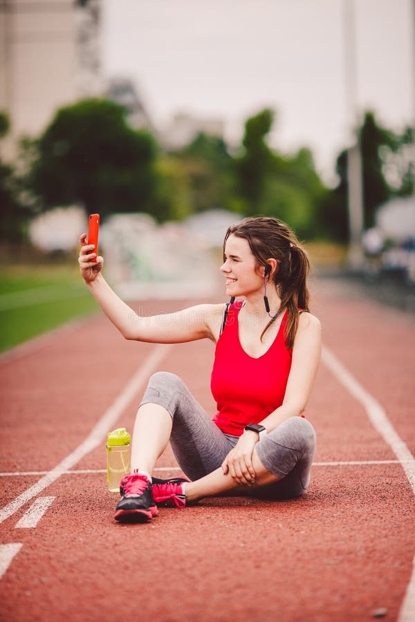 Atleta della giovane donna allo stile di vita di sport dello stadio, sedendosi sulla pista, prendente la foto del selfie su uno s fotografia stock libera da diritti Atleta della giovane donna allo stile di vita di sport dello stadio, sedendosi sulla pista, prendente la foto del selfie su uno s fotografia stock libera da diritti