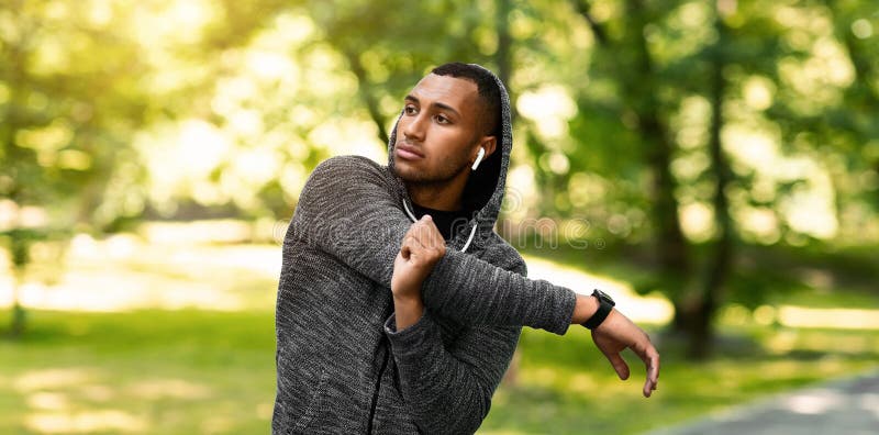 Atletico afroamericano a hoodie che allunga il braccio e ascolta musica dopo l'allenamento al parco estivo fotografia stock libera da diritti