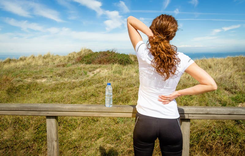 Back view of athletic young woman in sportswear touching her neck and lower back muscles by painful injury, over a nature background. Sport injuries concept. Back view of athletic young woman in sportswear touching her neck and lower back muscles by painful injury, over a nature background. Sport injuries concept.