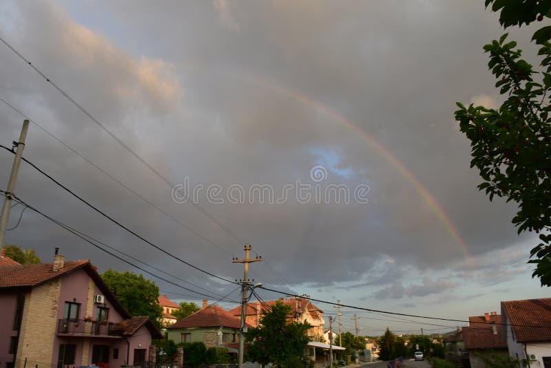 Houses in the village next to the road, and above them are gray clouds and rainbows across the clouds. Houses in the village next to the road, and above them are gray clouds and rainbows across the clouds