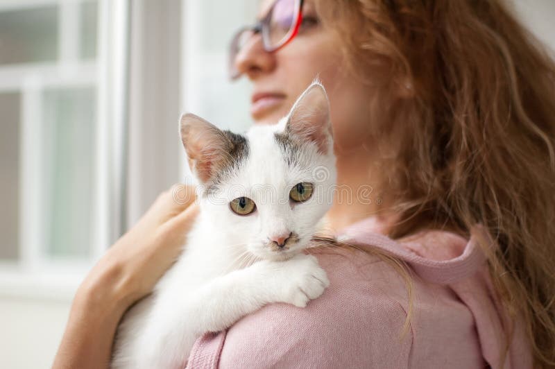 Attractive girl in pink clothes and eyeglasses with withe cat in her arms standing near the window at home. Pet owner concept. Attractive girl in pink clothes and eyeglasses with withe cat in her arms standing near the window at home. Pet owner concept.