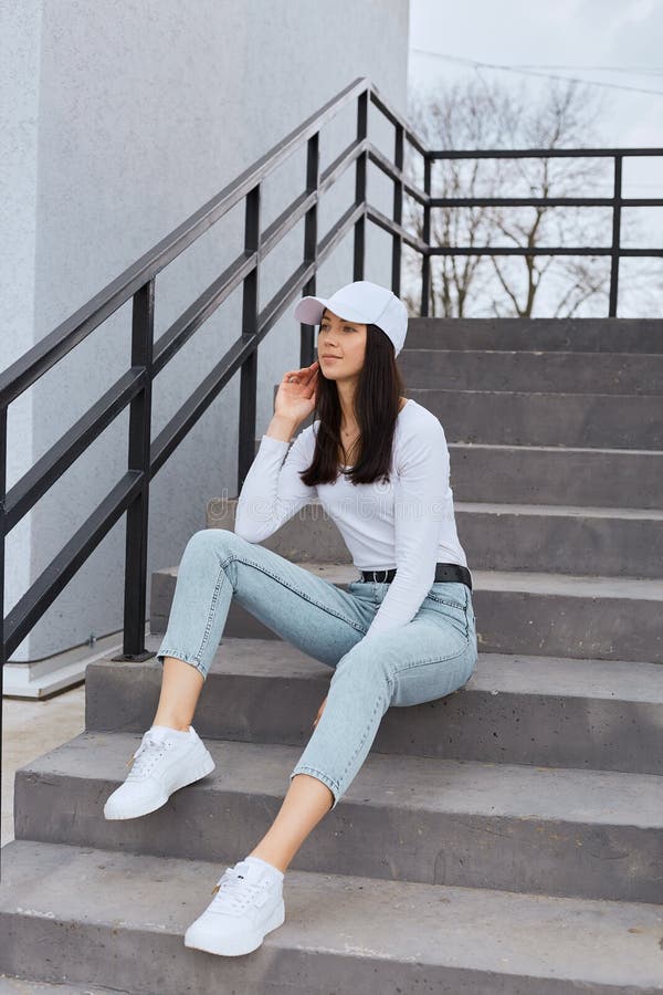 Atractiva Mujer Con Elegante Jeans Camisa Blanca Y Gorra De Béisbol Sentado  En Las Escaleras Y Mirando Hacia Otro Lado Con Reflexi Foto de archivo -  Imagen de gente, feliz: 216759084