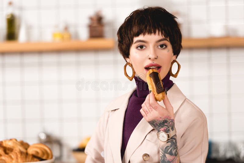 Atractiva mujer de negocios de moda degustando deliciosos eclair y mirando a la cámara. imagenes de archivo