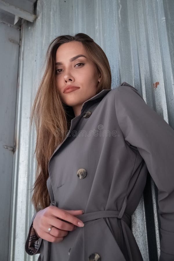 Atractivo modelo asiático joven con abrigo de trinchera gris parado cerca de la pared al aire libre foto de archivo