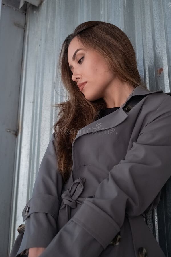 Atractivo modelo asiático joven con abrigo de trinchera gris parado cerca de la pared al aire libre fotos de archivo