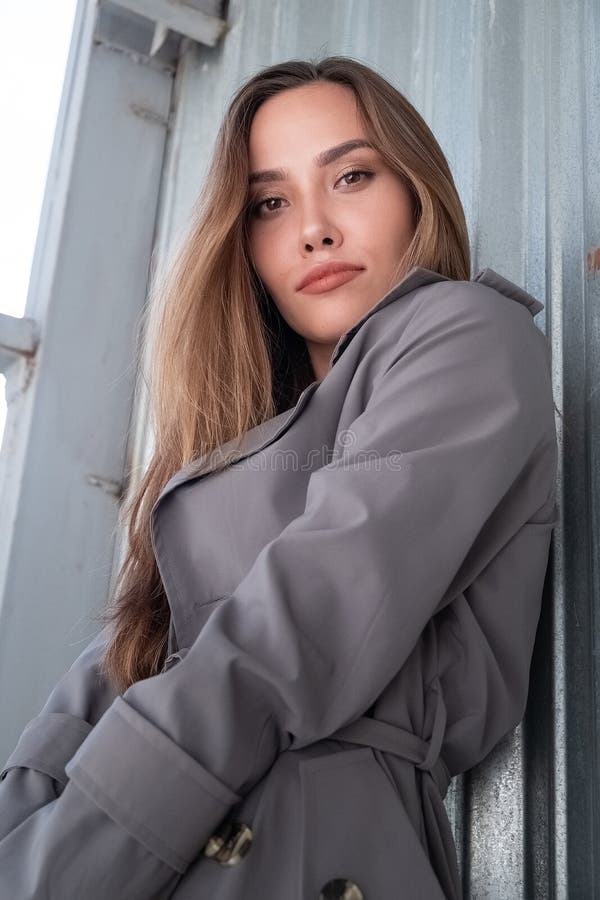 Atractivo modelo asiático joven con abrigo de trinchera gris parado cerca de la pared al aire libre foto de archivo