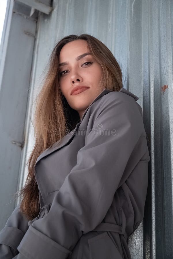 Atractivo modelo asiático joven con abrigo de trinchera gris parado cerca de la pared al aire libre fotos de archivo