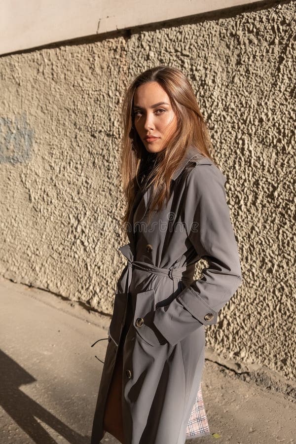 Atractivo modelo asiático joven con abrigo de trinchera gris parado cerca de la pared al aire libre foto de archivo