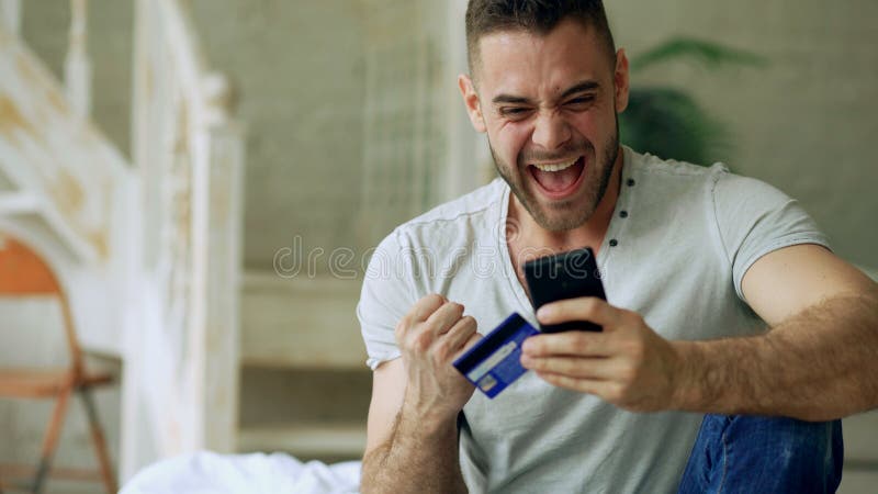 Atrakcyjny młody człowiek z smartphone i kredytowej karty zakupy na internecie siedzi na łóżku w domu obraz royalty free Atrakcyjny młody człowiek z smartphone i kredytowej karty zakupy na internecie siedzi na łóżku w domu obraz royalty free