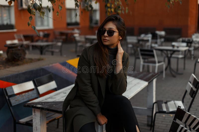 Atrativo e jovem mulher no revestimento verde à moda com os óculos de sol que sentam-se fora em um café Menina elegante imagem de stock royalty free Atrativo e jovem mulher no revestimento verde à moda com os óculos de sol que sentam-se fora em um café Menina elegante imagem de stock royalty free