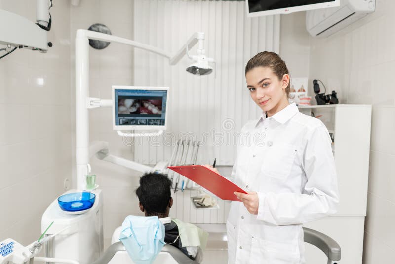 Att charma den härliga flickatandläkaren ser in i kameran Manlig patient för ung afrikansk amerikan på stol på den tand- kliniken fotografering för bildbyråer Att charma den härliga flickatandläkaren ser in i kameran Manlig patient för ung afrikansk amerikan på stol på den tand- kliniken fotografering för bildbyråer