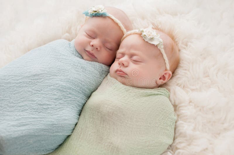 Seven week old fraternal, twin baby girls swaddled and sleeping on a white flokati rug. One sister is smiling. Seven week old fraternal, twin baby girls swaddled and sleeping on a white flokati rug. One sister is smiling.