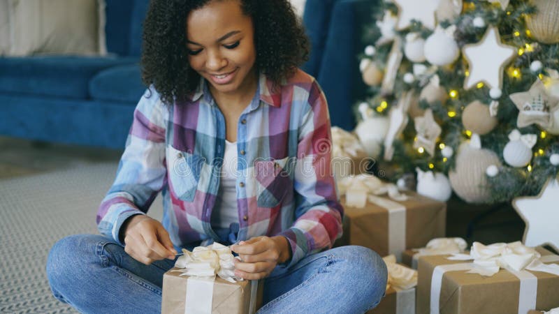 Attractive mixed race girl packing gift box near Christmas tree at home stock photo Attractive mixed race girl packing gift box near Christmas tree at home stock photo