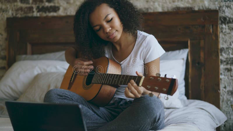 Attractive african american teenager girl concentraing learning to play guitar using laptop computer sitting on bed at royalty free stock photos Attractive african american teenager girl concentraing learning to play guitar using laptop computer sitting on bed at royalty free stock photos
