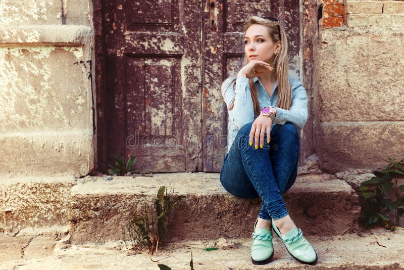 Attractive beautiful gentle girl sits in the city on the steps of the old building in jeans and fashion footwear