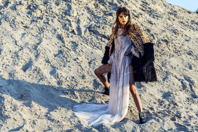 Attractive brunette woman in translucent beach long cover up with leopard fur coat posing on sandy beach at sunset. standing and royalty free stock image Attractive brunette woman in translucent beach long cover up with leopard fur coat posing on sandy beach at sunset. standing and royalty free stock image